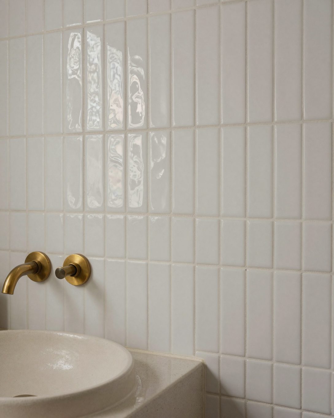 Illustration: close-up of a bathroom wall clad in small handmade glossy white zellige tiles with uneven wavy surfaces and slightly irregular edges catching the light, above a microcement vanity with a handmade cream ceramic basin and a warm brass wall tap